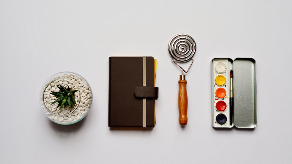 Uma composição minimalista com uma pequena planta suculenta, um caderno elegante, um item de cozinha em espiral e uma caixa de aquarela, representando diversas opções de presentes criativos e funcionais.